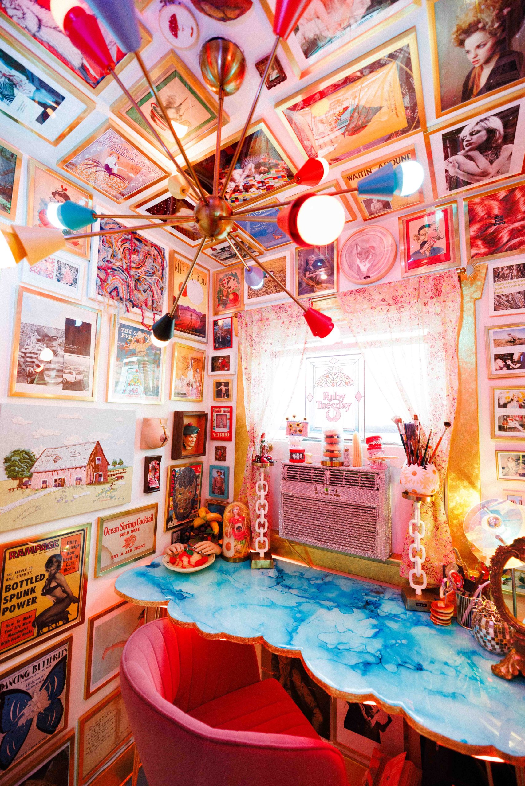 An art-filled room with a blue cloud-patterned desk, pink chair, and colorful branching ceiling light.