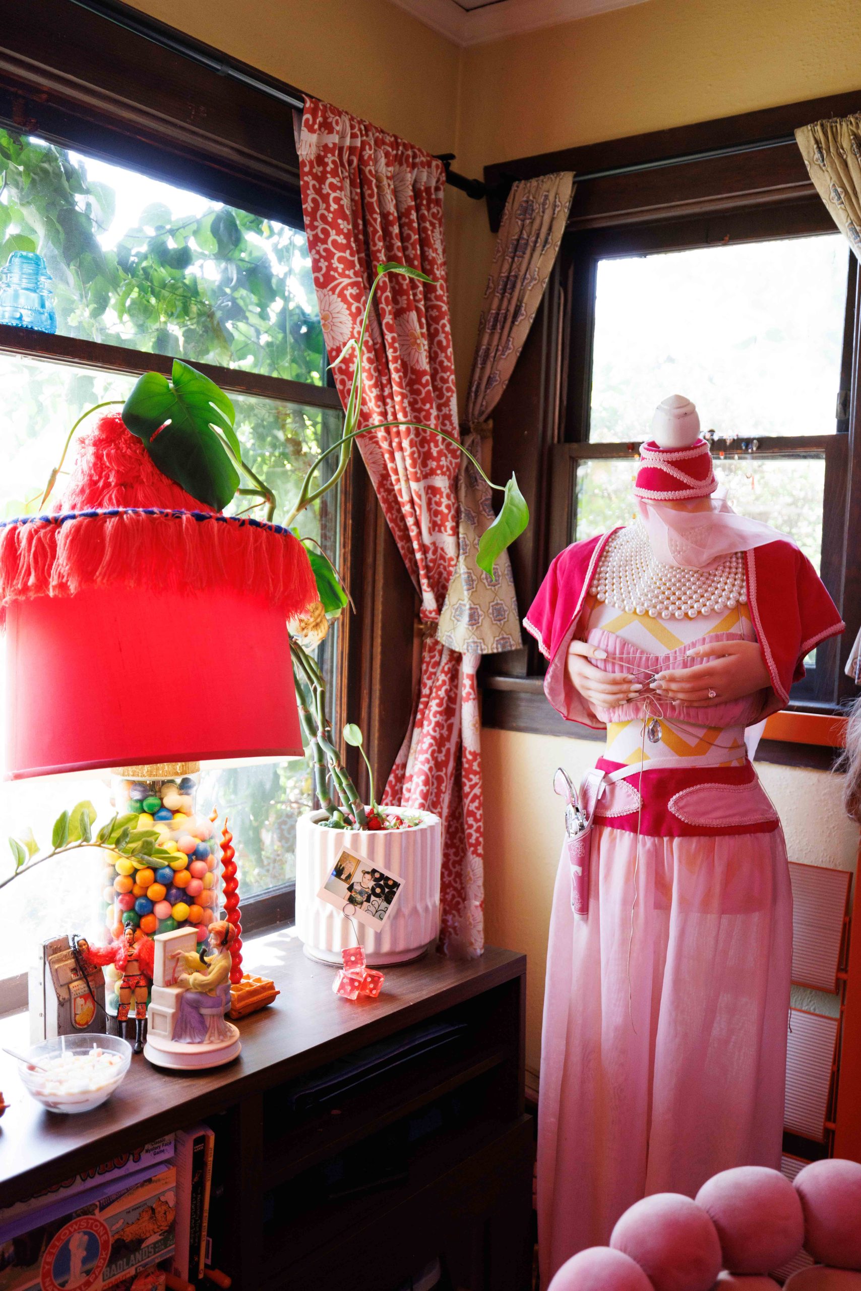 A mannequin in a pink dress stands beside a window and a red lampshade.