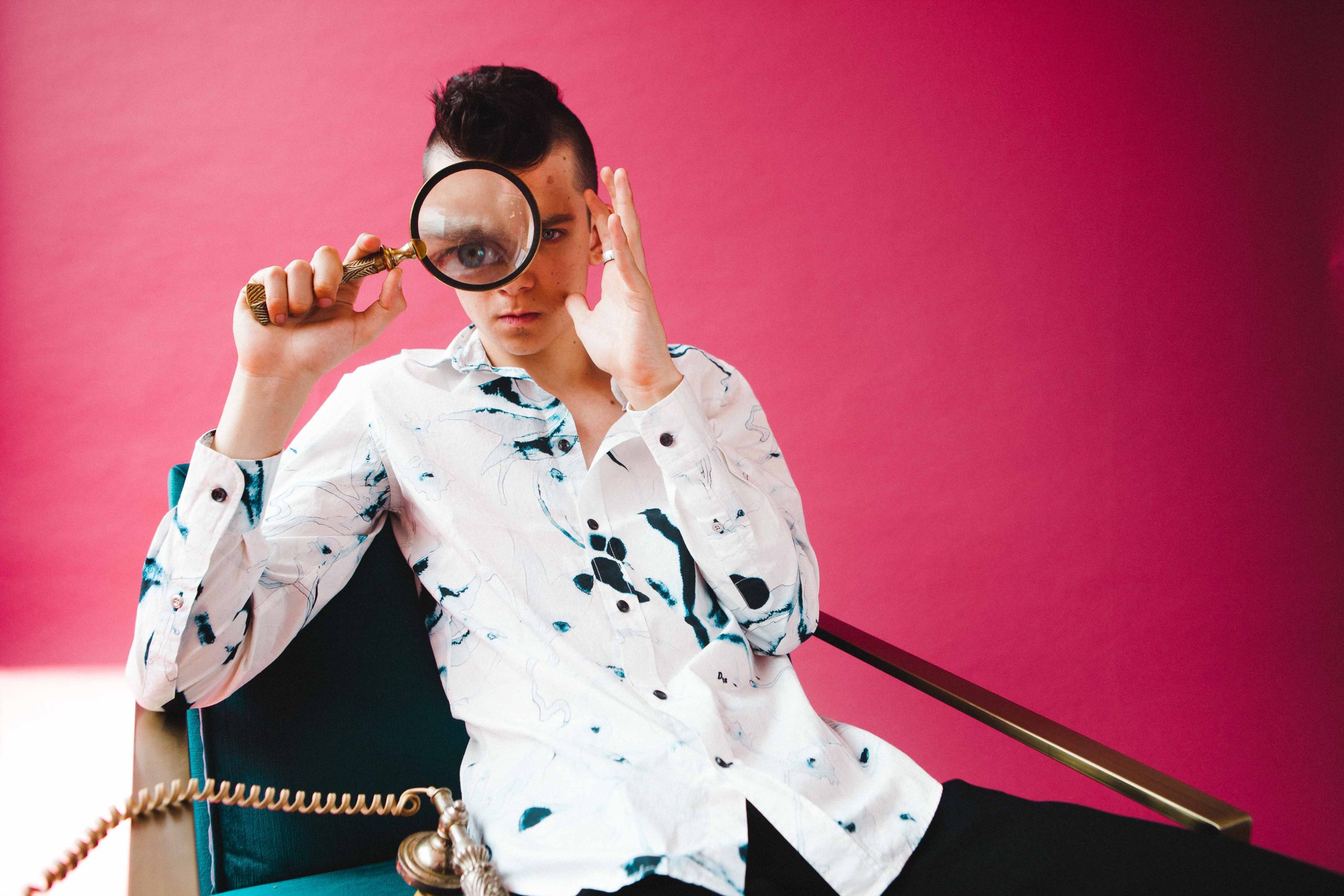 Asa Butterfield holds a magnifying glass over one eye against a vivid pink background.