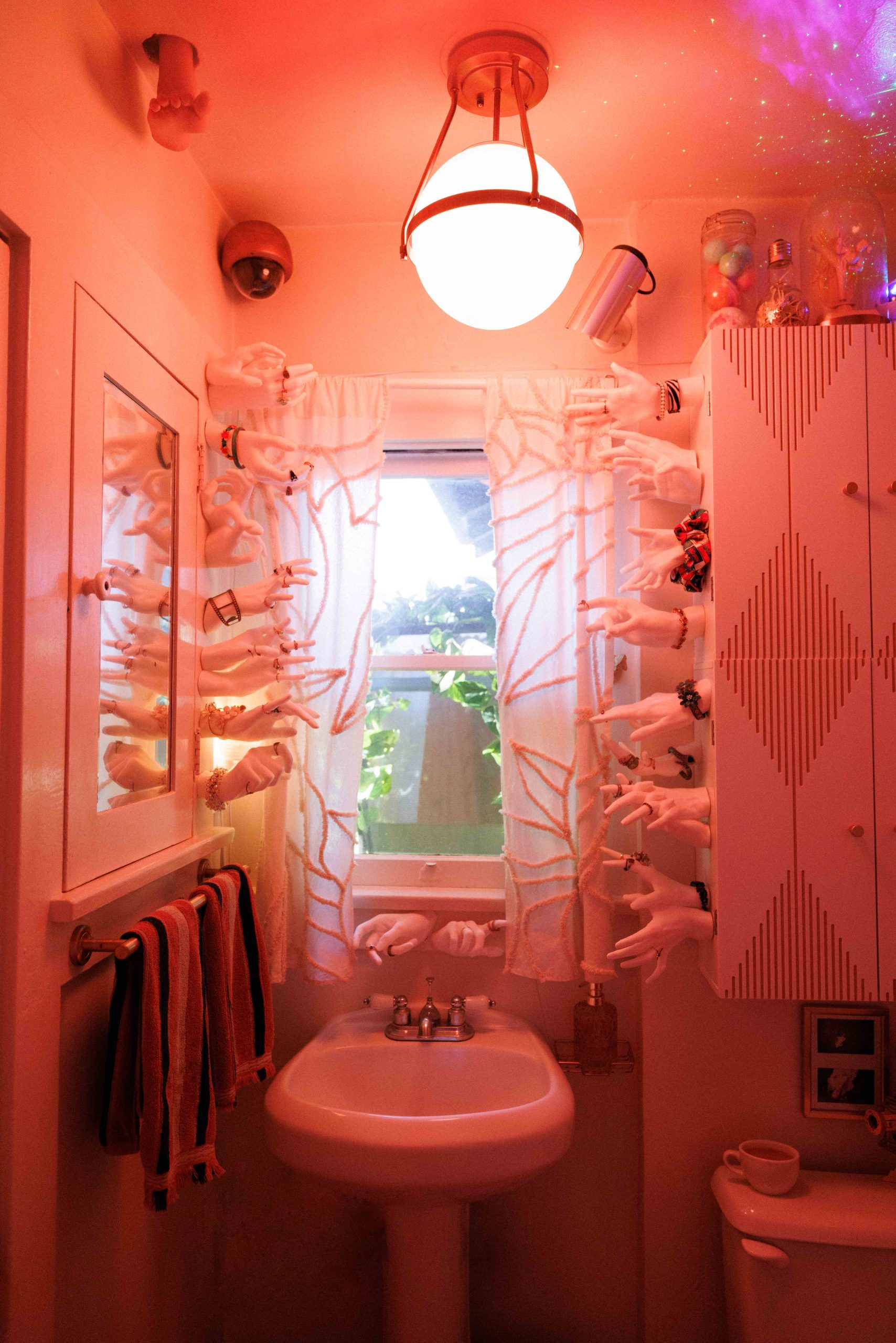 Bathroom lit in red, with security cameras and rows of mannequin hands around the sink and window.
