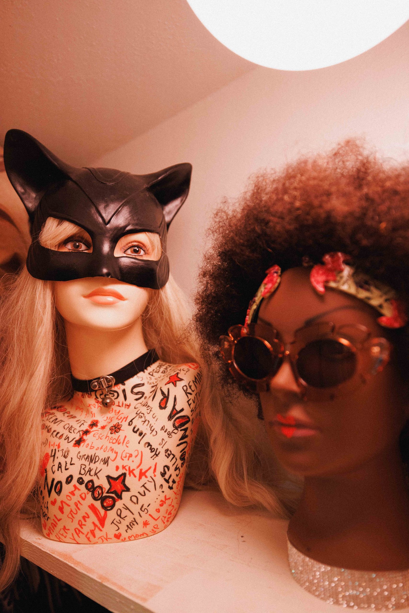 Two mannequin heads, one in a black cat mask and the other wearing sunglasses and a curly wig.