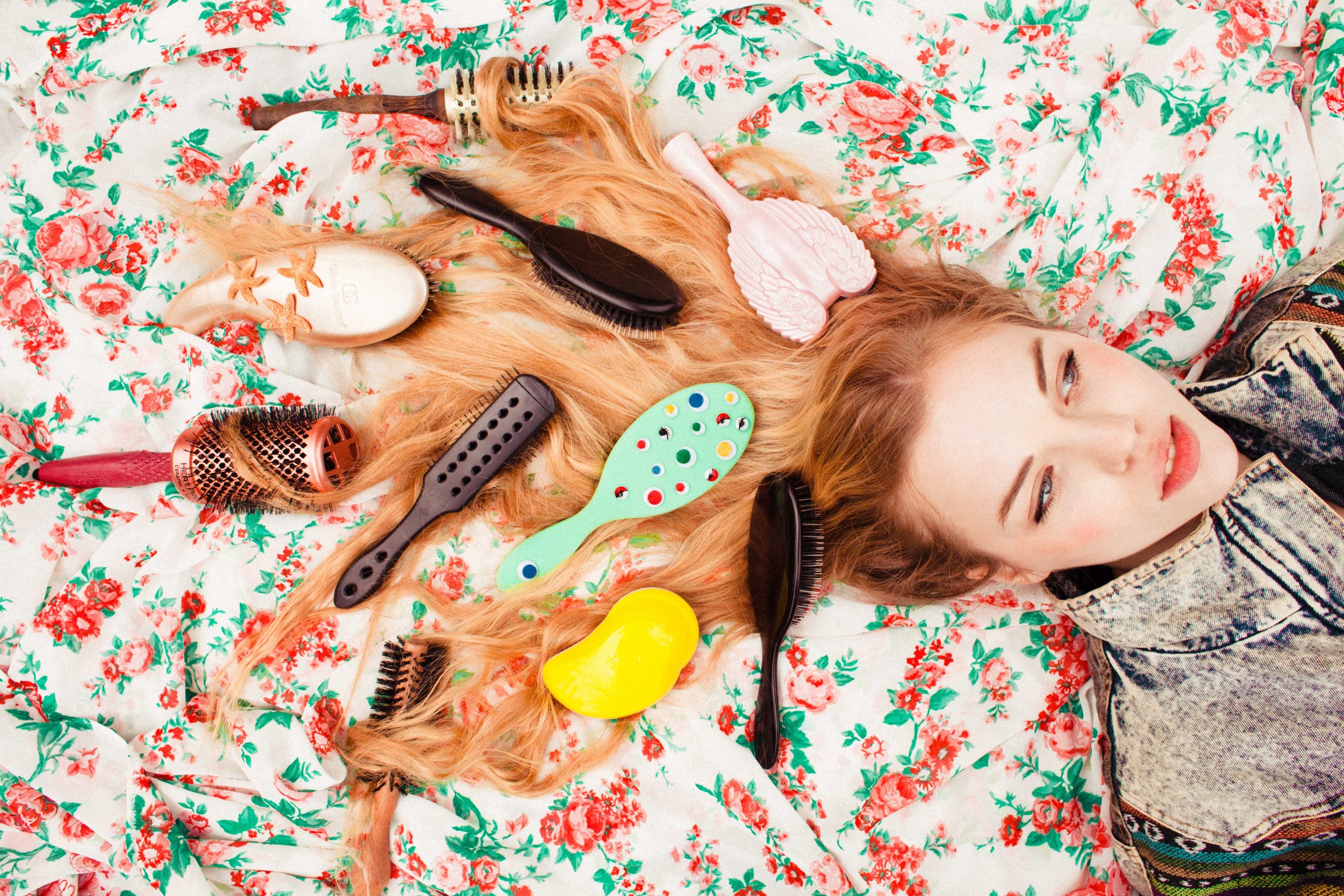 Ruby June lies on floral fabric with colorful hairbrushes arranged around her hair.