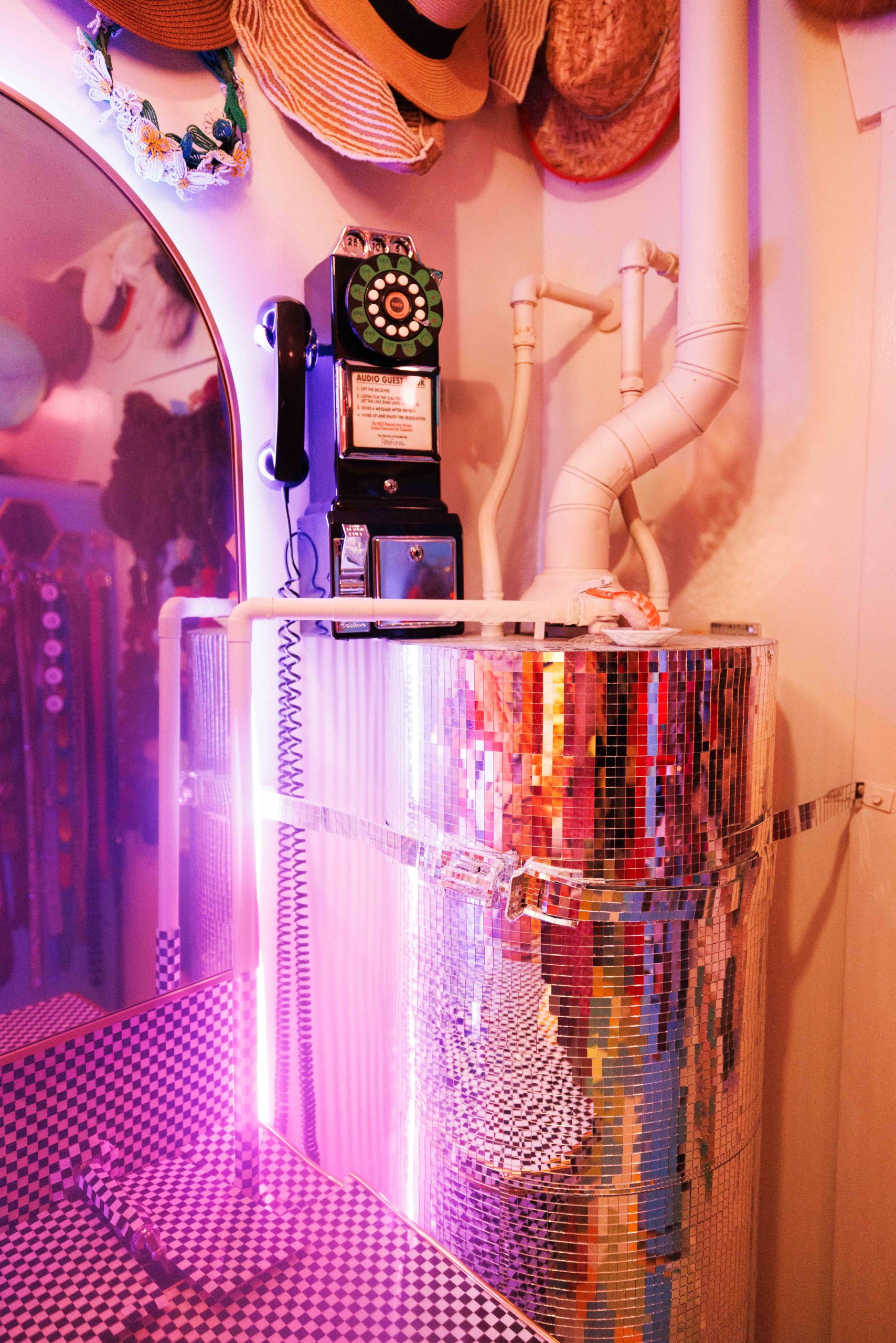 A black rotary telephone labeled Audio Guide sits on a mirrored mosaic cylinder beneath a collection of hats.