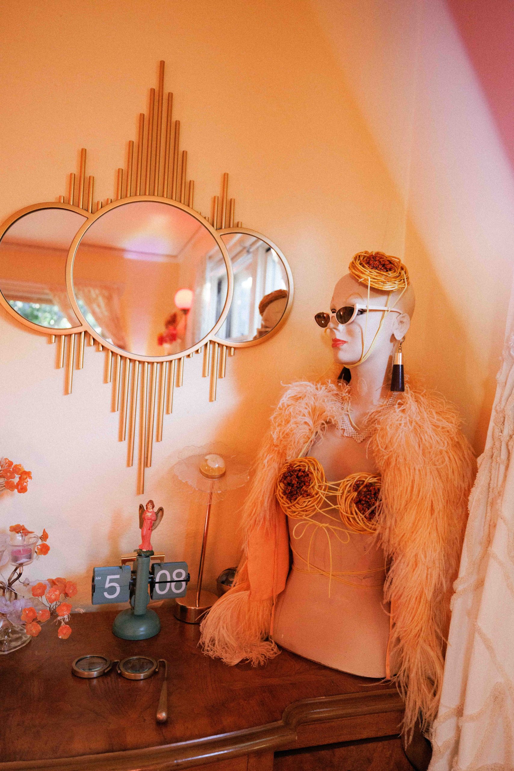 A mannequin wearing sunglasses and a feathered garment beside round mirrors and a dresser.