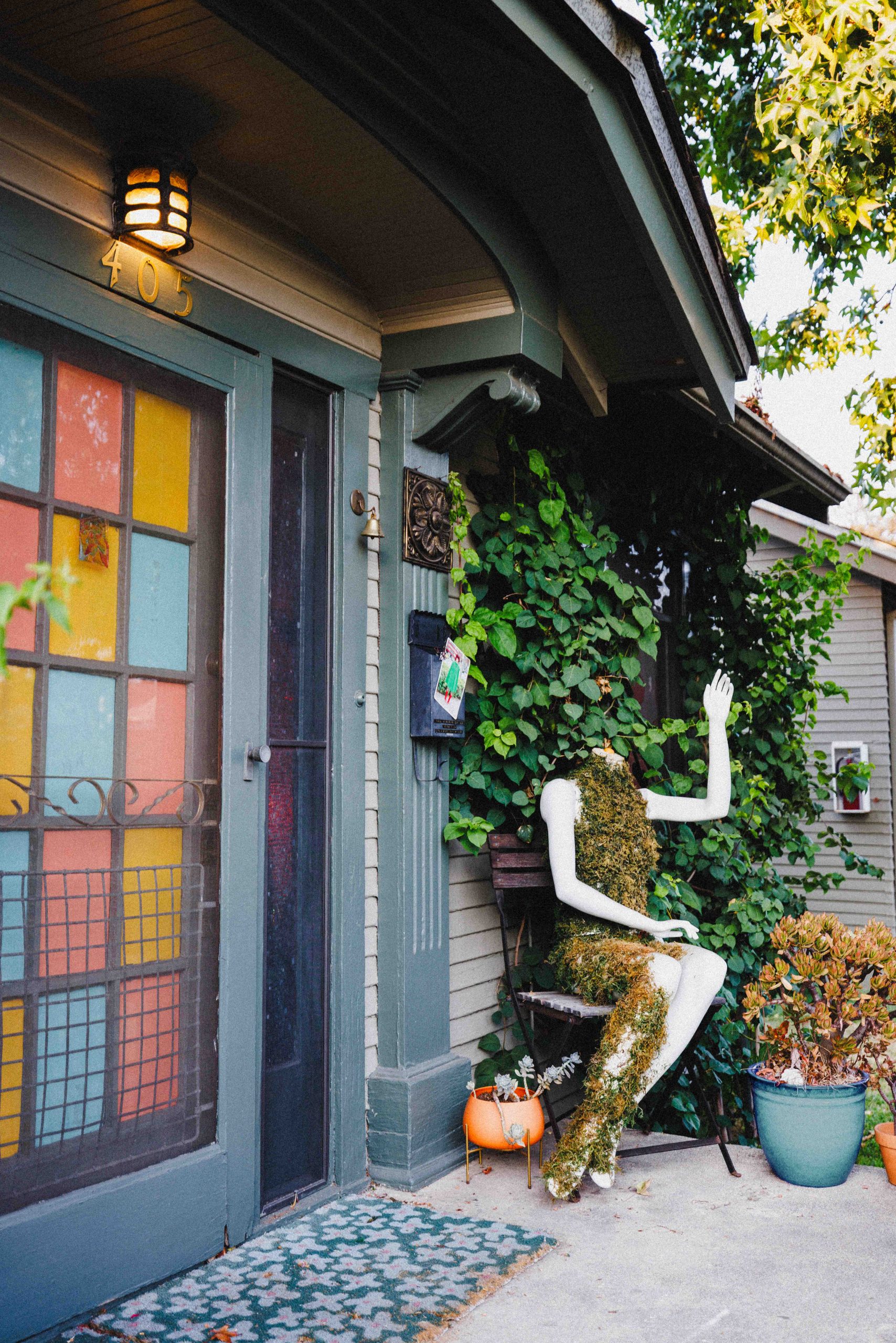 A seated mannequin figure beside the Museum’s colored-glass entrance, surrounded by plants.