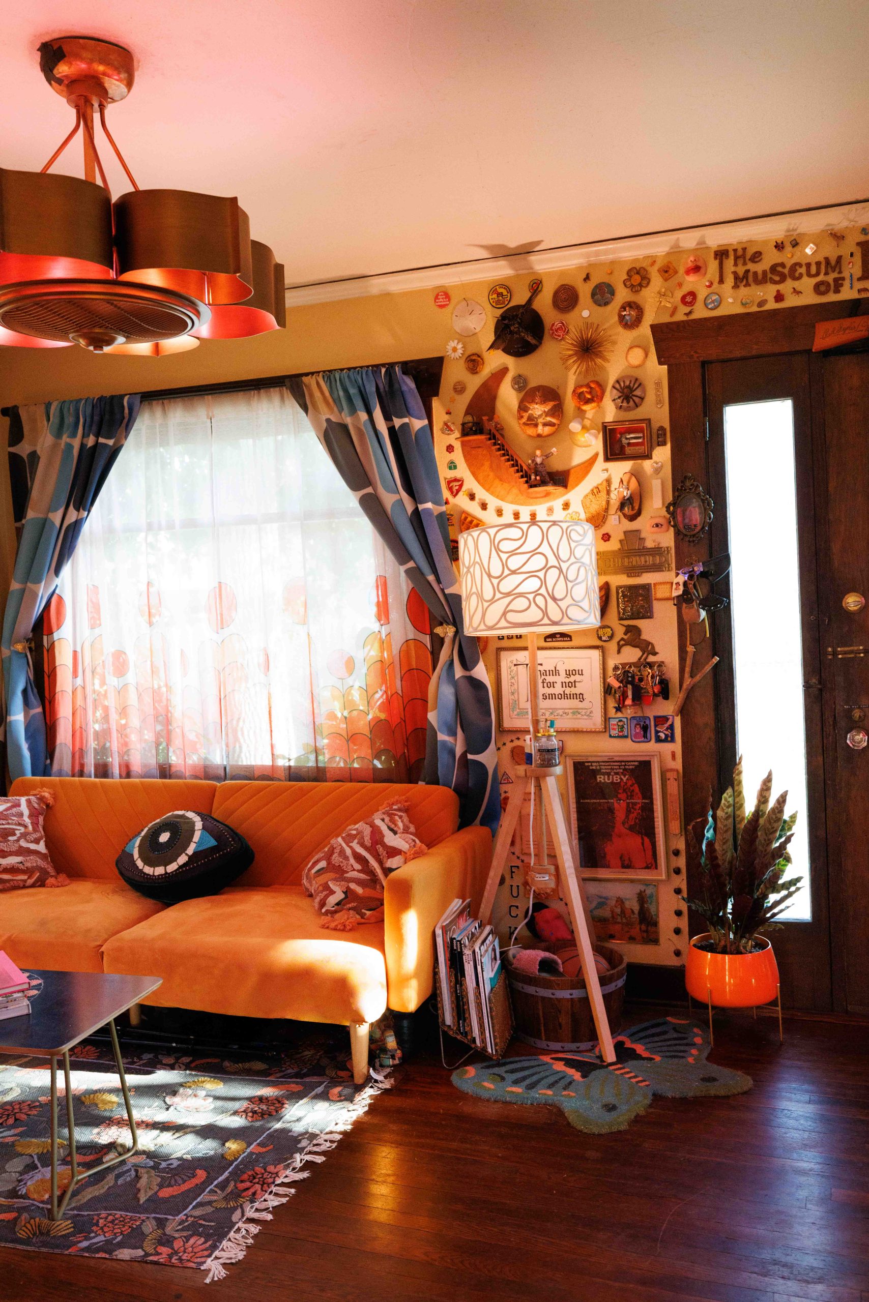 An orange sofa and glass coffee table in a room filled with patterned curtains, artwork, and objects.