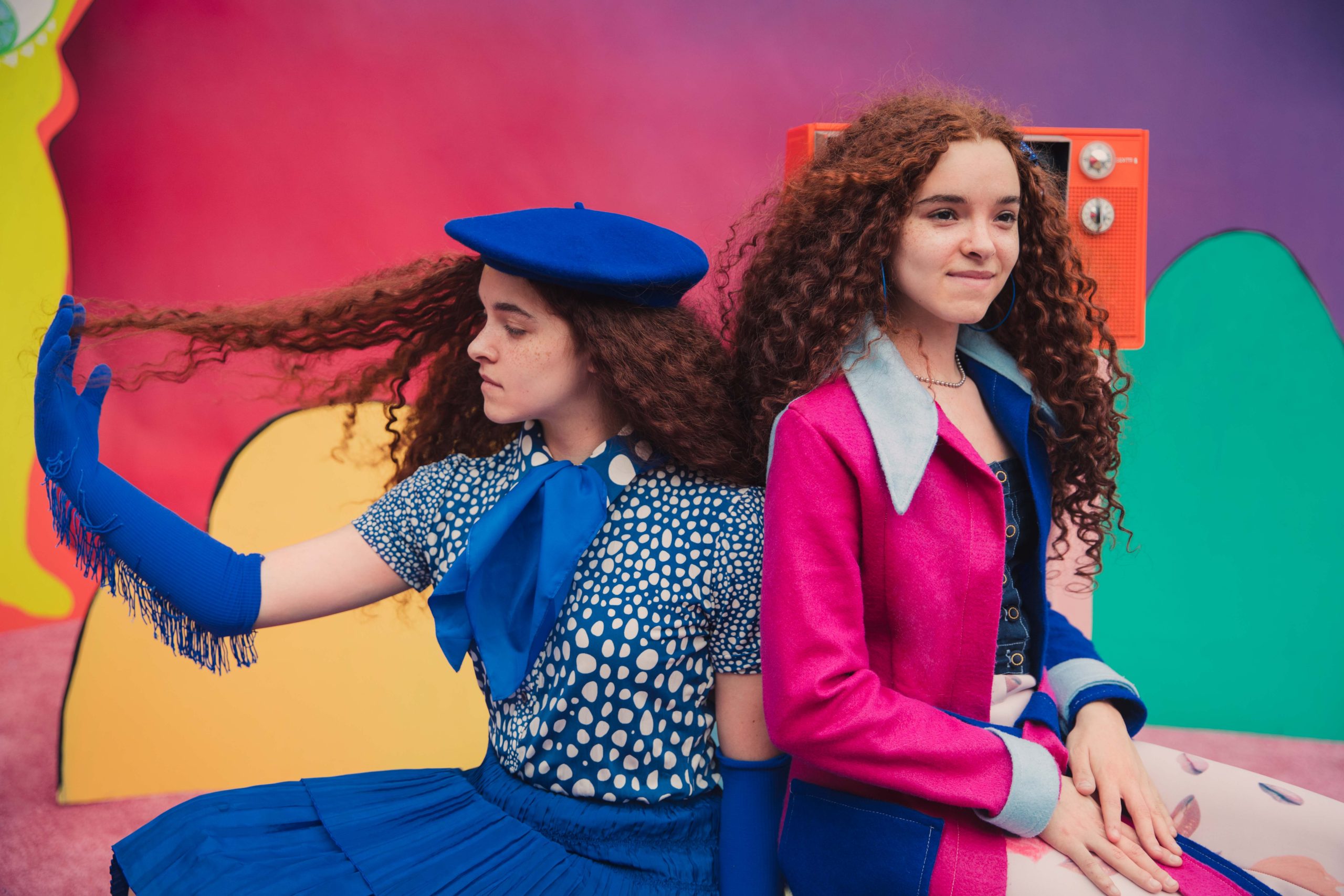 Two models in blue and pink sit back to back against a brightly colored studio backdrop.