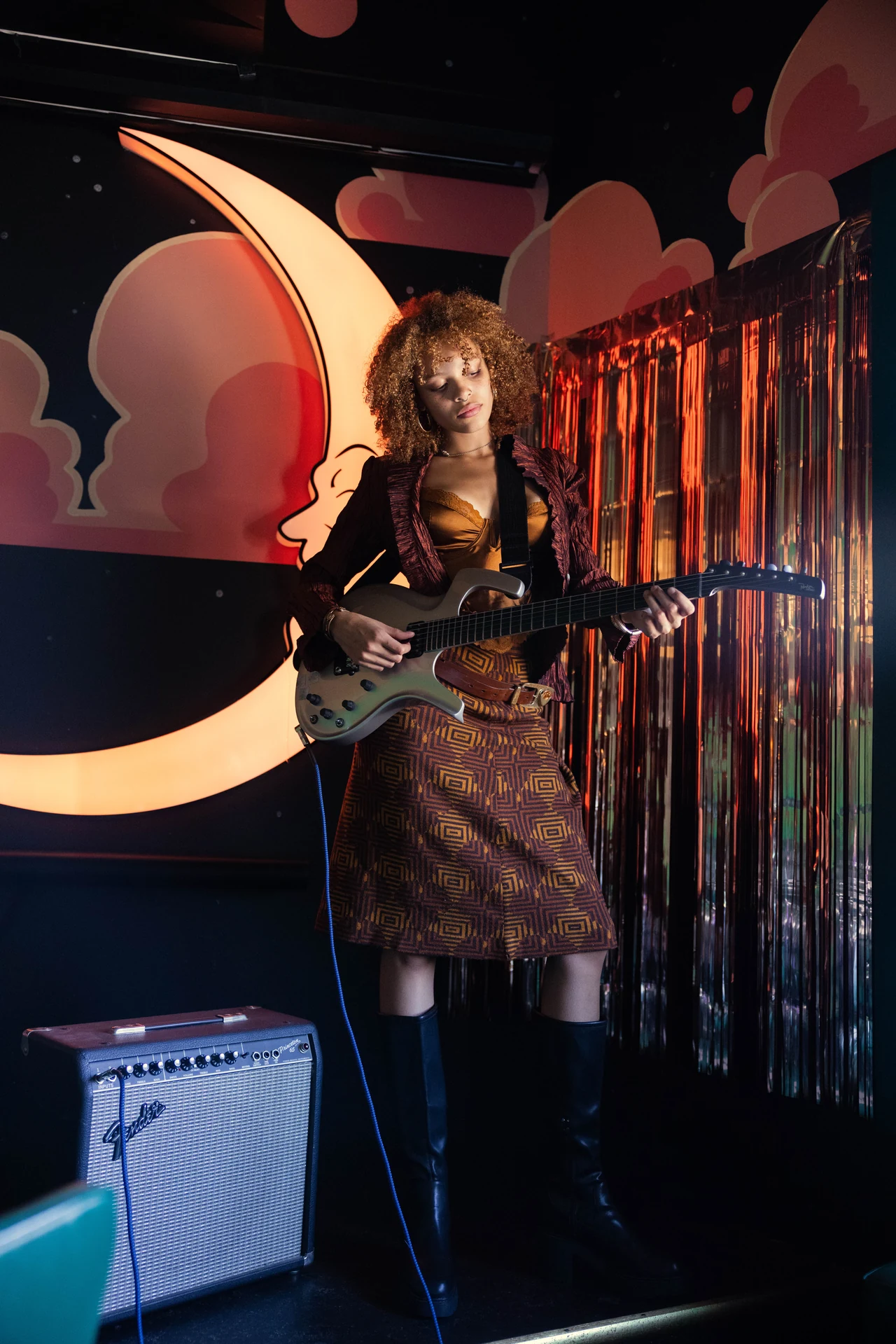 Full-length portrait wearing the guitar-pick belt and holding an electric guitar beside an amplifier.