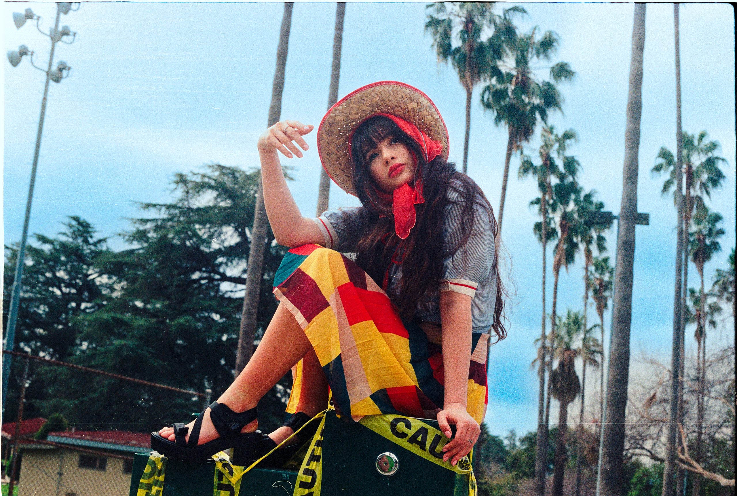 Malina Weissman sits on a skateboard outdoors, wearing a straw hat, with palm trees behind her.